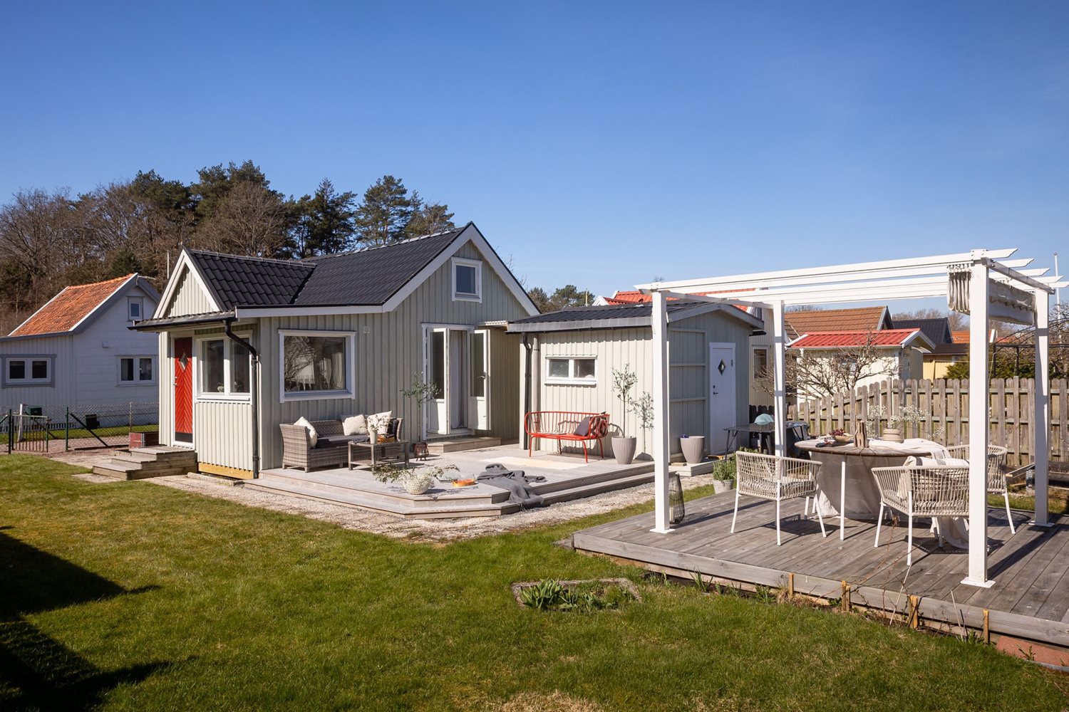 Vy inifrån trädgård mot stugan och uteplatsen och pergolan